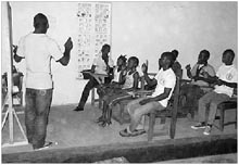 Afrikanische gehörlose Schüler sitzen auf Stühlen im Unterrichtsraum; der Lehrer erklärt an der Tafel etwas.