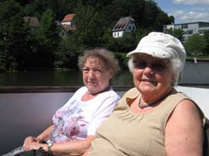Zwei Frauen sitzen gemütlich an Deck des Neckarschiffes; im Hintergrund das Wasser und Häuser am grünen Ufer