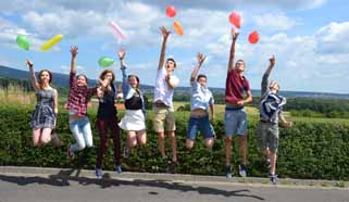 Acht Jugendliche springen in die Luft und greifen nach Luftballons; im Hintergrund grüne Wiese und blauer Himmel